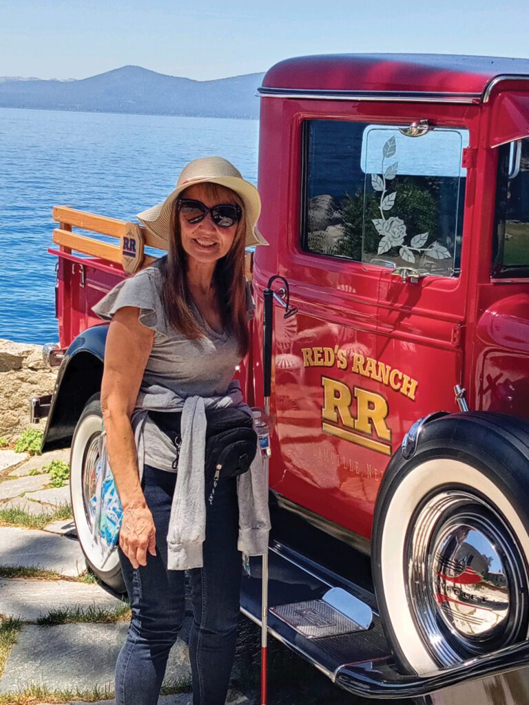 Mrs WH stands with her red tipped white cane near a vintage truck on the beach smiling